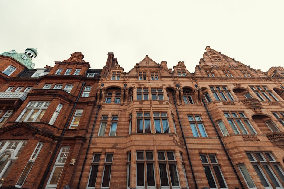 A low-angle view of a historic multi-story building in Knightsbridge, featuring ornate Victorian-style architecture with detailed brickwork, arched windows, and decorative stone elements. The building’s facade is constructed from red and brown bricks with contrasting stone accents around the windows and roofline. The upper floors include large windows, some with small balconies, and the roof has a distinctive turret and spire visible against the cloudy sky. In front, a paved pavement area is visible, possibly an entryway or loading zone used during house removals. This scene depicts the exterior environment where Man and Van Knightsbridge may facilitate furniture transport and home relocations, capturing the process of moving large furniture and boxes near the building’s entrance, with a focus on loading and packing activities as part of a professional removals operation.
