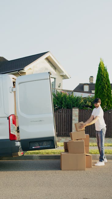 A man wearing a white t-shirt and jeans is standing beside an open white commercial van on a residential street, preparing to load three cardboard boxes onto the vehicle. The boxes are stacked on the ground, and the man is holding a small cardboard box, ready to place it inside the van. The background features a white house with a grey roof, a black fence, and tall green trees, under a clear sky with natural daylight. This scene depicts the process of home relocation, with the man and van from Man and Van Knightsbridge involved in furniture transport and packing and moving activities, highlighting the logistical steps of small flat moves in the Brompton Road and Knightsbridge area.