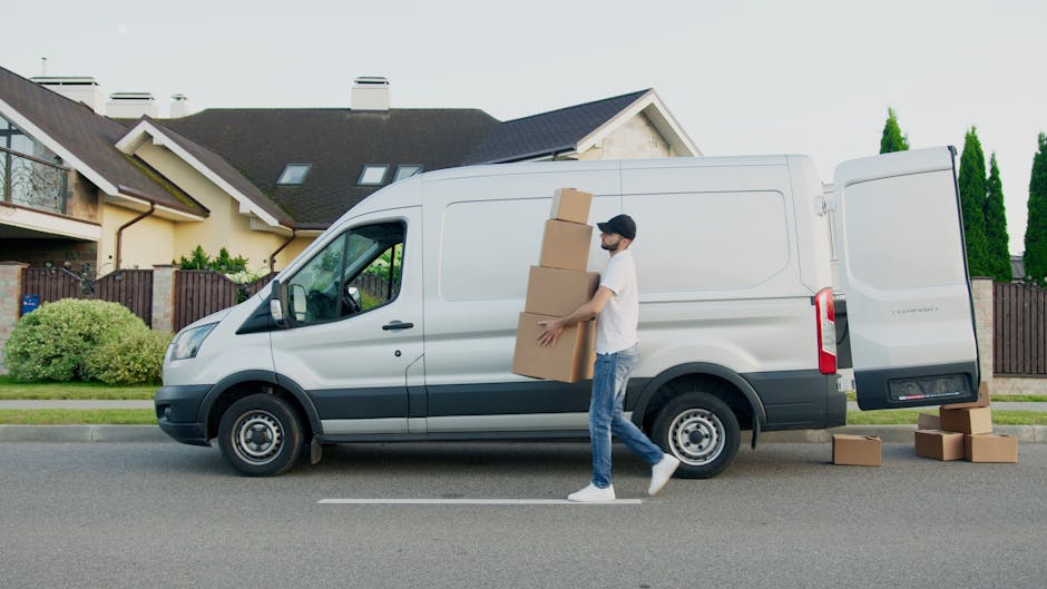 A man wearing a white t-shirt and jeans is standing beside an open white commercial van on a residential street, preparing to load three cardboard boxes onto the vehicle. The boxes are stacked on the ground, and the man is holding a small cardboard box, ready to place it inside the van. The background features a white house with a grey roof, a black fence, and tall green trees, under a clear sky with natural daylight. This scene depicts the process of home relocation, with the man and van from Man and Van Knightsbridge involved in furniture transport and packing and moving activities, highlighting the logistical steps of small flat moves in the Brompton Road and Knightsbridge area.