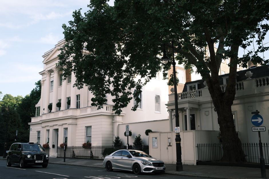Photograph of a white, multi-story building with classical architectural features, including decorative columns and balustrades, situated on a tree-lined street. In the foreground, there are two parked vehicles: a black van and a silver sedan. The scene is captured in daylight with sunlight illuminating the building's facade, and a large tree with dense green foliage partially obscures the upper part of the building. A street lamp and traffic signs, including a one-way sign, are visible near the curb. Given the context of house removals and moving logistics, this image appears to depict the exterior environment where furniture transport and packing activities could take place, with Man and Van Knightsbridge potentially involved in the home relocation process outside this property.