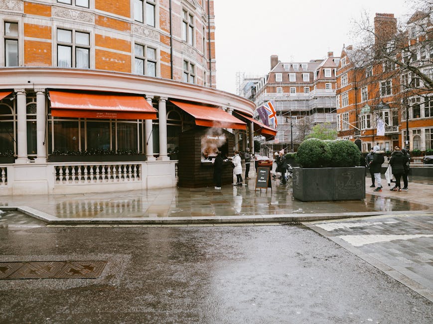 A street scene outside a building with red brick and cream-colored architectural details on a rainy day in Knightsbridge. Several people are gathered around a small outdoor area with a black charcoal grill emitting smoke, near a storefront with orange awnings. The pavement is wet, reflecting the overcast sky, and there is a large grey planter with greenery on the sidewalk. In the background, multi-storey residential buildings and scaffolding are visible, along with a Union Jack flag. A man and a woman are seen carrying boxes and preparing for a home relocation or furniture transport process, supported by a professional removals service such as Man and Van Knightsbridge. Movable items like cardboard boxes, wooden crates, and packaging materials are being handled, with some placed near a van parked nearby for loading or unloading. The scene captures the logistical activity involved in packing and moving belongings in an urban environment, aligned with council rules for removals in the Knightsbridge area.