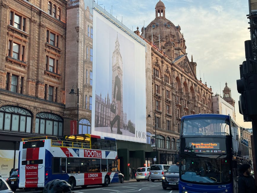 A busy street scene in Knightsbridge featuring historic brick buildings with ornate architectural details and large windows. In the foreground, a double-decker bus displaying a route to Kensington is parked alongside a red and white tour bus branded 'TOOTbus', both situated on the road. The street is filled with several parked cars, and a few pedestrians are visible on the pavement, some wearing face masks. On the building façade, a large vertical billboard advertising Dior is visible, depicting a model in a black outfit carrying a bag, standing in front of the UK Houses of Parliament with Big Ben in the background. The scene is illuminated by natural daylight, with a clear sky overhead, reflecting a typical city environment relevant to house removals or moving services in the area around Harrods, with architecture and street elements capturing the context of urban transit and logistics.
