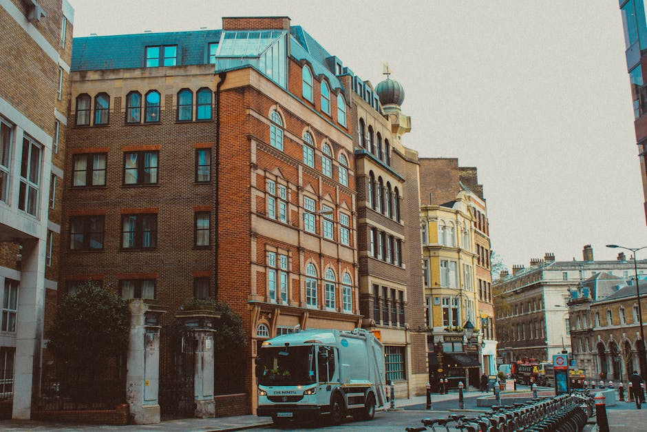 A street scene in Knightsbridge showing historic multi-storey buildings with a mix of brick and ornate facades, some featuring arched windows and decorative stonework, under a light overcast sky. A blue and white van belonging to Man and Van Knightsbridge is parked on the pavement near a gated entrance, which is surrounded by a low stone wall and iron gates. Several cardboard boxes, wrapped in clear plastic and brown packing tape, are stacked on the pavement, ready for home relocation or furniture transport. A moving trolley and lifting straps are visible, indicating active loading or unloading, with some boxes being carried by movers inside the building entrance. The street includes bollards and bike racks, with a few pedestrians and vehicles in the background, illustrating typical urban moving logistics near Harrods and central London. This scene captures the typical environment for house removals involving packing, transporting, and loading process supported by local professional services.
