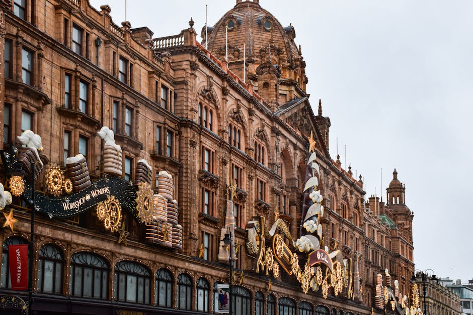 A busy street scene in Knightsbridge featuring historic brick buildings with ornate architectural details and large windows. In the foreground, a double-decker bus displaying a route to Kensington is parked alongside a red and white tour bus branded 'TOOTbus', both situated on the road. The street is filled with several parked cars, and a few pedestrians are visible on the pavement, some wearing face masks. On the building façade, a large vertical billboard advertising Dior is visible, depicting a model in a black outfit carrying a bag, standing in front of the UK Houses of Parliament with Big Ben in the background. The scene is illuminated by natural daylight, with a clear sky overhead, reflecting a typical city environment relevant to house removals or moving services in the area around Harrods, with architecture and street elements capturing the context of urban transit and logistics.