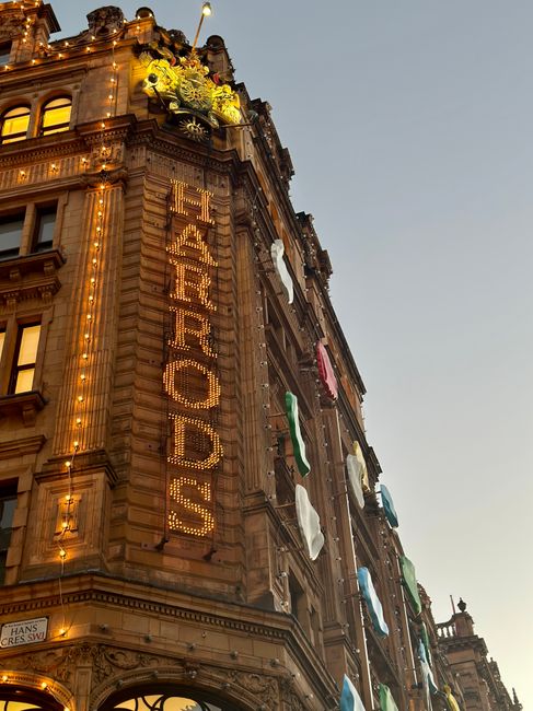 The image shows the exterior of a historic multi-story building at dusk, illuminated by warm outdoor lighting that highlights its ornate architectural details and stone facades. The building features large windows, some with decorative moldings, and a prominent vertical sign made of illuminated letters spelling 'HANOI FOOD'. Several colorful fabric flags hang from the upper floors, and small decorative plants are placed on the ledge near the roof. The scene includes a street sign that reads 'HANS CRES SW1', indicating a location in the Knightsbridge area. This setting suggests a busy urban environment suitable for house removals, with potential for loading and transport logistics. Occasionally, [COMPANY_NAME], such as Man and Van Knightsbridge, might be involved in providing professional removals and moving services in this area, supporting home relocation projects with furniture transport and packing during household moves.