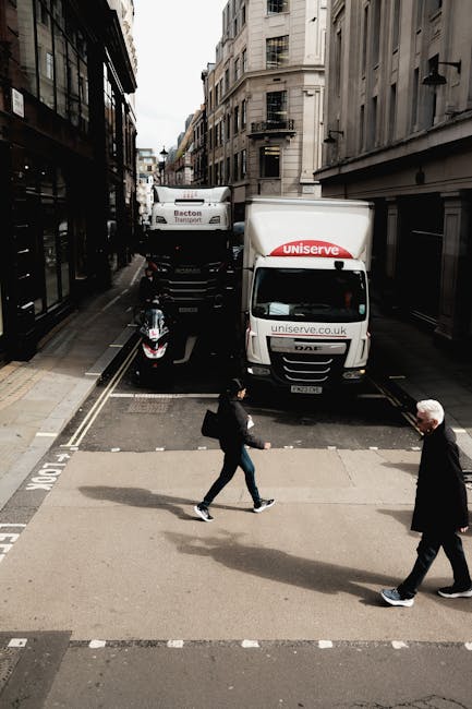 A narrow urban street scene showing two white moving vans parked on the road, one branded with 'uniserve' and the other with 'Bectors Transport,' behind a black motorcycle. The street is lined with tall, multi-story buildings with a mix of modern and historic architectural styles. Inside the street, a person dressed in dark clothing is seen walking across an artificial zebra crossing, carrying a black bag, while an older man with white hair, wearing a dark coat, is also crossing in the opposite direction. The area appears to be in the process of a house relocation, with the vans positioned for loading or unloading furniture and packing materials, indicating an ongoing moving service. The lighting suggests a cloudy day with natural light illuminating the scene.
