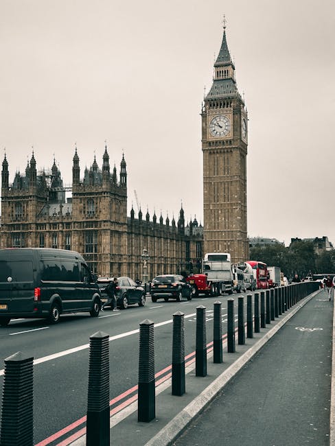 A scene of a street in London with the iconic Elizabeth Tower, commonly known as Big Ben, visible in the background. The street features several vehicles, including black vans, white trucks, and red double-decker buses, parked along the curb, which is lined with black bollards. The road is divided by a white dashed line, indicating multiple lanes of traffic. In the foreground, there is a pedestrian walkway separated from the roadway by a curb, and the pavement is visible. The overall scene is set during daytime with overcast skies, and the setting reflects a busy urban environment suitable for home relocation or furniture transport services, with [COMPANY_NAME] occasionally mentioned for context of moving logistics.