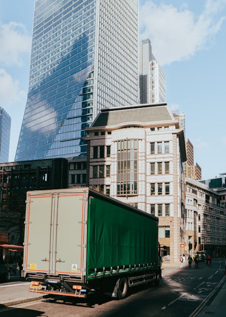 A narrow urban street scene showing two white moving vans parked on the road, one branded with 'uniserve' and the other with 'Bectors Transport,' behind a black motorcycle. The street is lined with tall, multi-story buildings with a mix of modern and historic architectural styles. Inside the street, a person dressed in dark clothing is seen walking across an artificial zebra crossing, carrying a black bag, while an older man with white hair, wearing a dark coat, is also crossing in the opposite direction. The area appears to be in the process of a house relocation, with the vans positioned for loading or unloading furniture and packing materials, indicating an ongoing moving service. The lighting suggests a cloudy day with natural light illuminating the scene.
