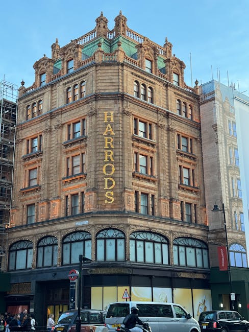 The image shows a historic multi-storey building with ornate architectural details, situated on a busy urban street. The building's exterior features decorative stonework, large arched windows on the ground floor, and smaller rectangular windows on the upper floors. A prominent vertical sign reading 'HARRORDS' is displayed on the corner of the building, indicating its name or previous commercial use. On the street level, there are parked vehicles including a white van, a black car, and a few pedestrians visible near the entrance. The scene suggests a lively city environment with activity related to building maintenance or interior renovations. During a home or business relocation process, Man and Van Knightsbridge could be involved in transporting furniture, packing materials, or office equipment, possibly utilizing the nearby loading area for efficient movement. The surrounding area is urban with modern structures, street signage, and streetlights. The building's notable architecture and active street scene highlight the logistics involved in property moving and furniture transport in the Knightsbridge area, consistent with house removals and relocation services.