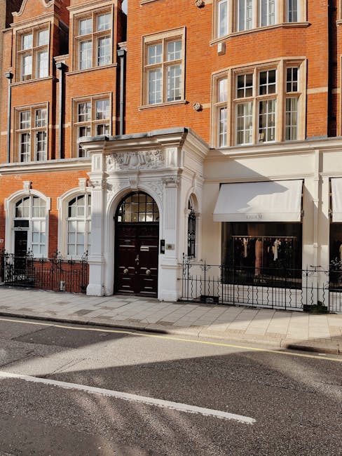A street view of a historic townhouse with a red brick façade and large sash windows, situated in Sloane Street near Knightsbridge; the ground floor features an ornate white stone entrance with decorative carvings and a black wooden door, bordered by black wrought iron fencing. To the right of the entrance, there is a shop window with a white awning, displaying clothing or merchandise inside. The pavement is clean and concrete, with a few cardboard boxes and protective furniture pads visible near the doorway, indicating recent or ongoing house removals. In the foreground, a black van with open rear doors is parked on the street, partially loaded or prepared for furniture transport, with visible trolleys and packing materials nearby, supporting the context of professional removals and home relocation services provided by Man and Van Knightsbridge.