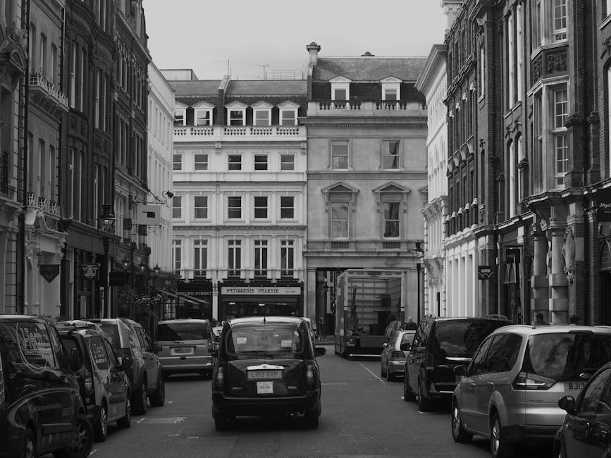 The image shows a historic multi-storey building with ornate architectural details, situated on a busy urban street. The building's exterior features decorative stonework, large arched windows on the ground floor, and smaller rectangular windows on the upper floors. A prominent vertical sign reading 'HARRORDS' is displayed on the corner of the building, indicating its name or previous commercial use. On the street level, there are parked vehicles including a white van, a black car, and a few pedestrians visible near the entrance. The scene suggests a lively city environment with activity related to building maintenance or interior renovations. During a home or business relocation process, Man and Van Knightsbridge could be involved in transporting furniture, packing materials, or office equipment, possibly utilizing the nearby loading area for efficient movement. The surrounding area is urban with modern structures, street signage, and streetlights. The building's notable architecture and active street scene highlight the logistics involved in property moving and furniture transport in the Knightsbridge area, consistent with house removals and relocation services.
